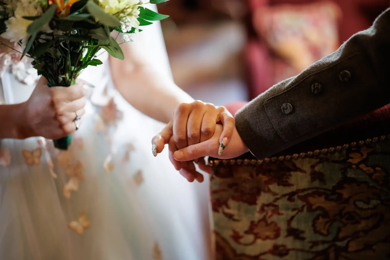 A bride holding a bouquet clasps hands with another person in formal attire, both standing close together in an indoor setting.