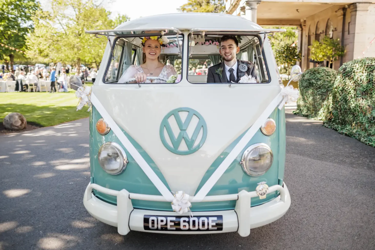 A bride and groom sit in the front seats of a vintage mint green and white Volkswagen van decorated with ribbons and flowers.