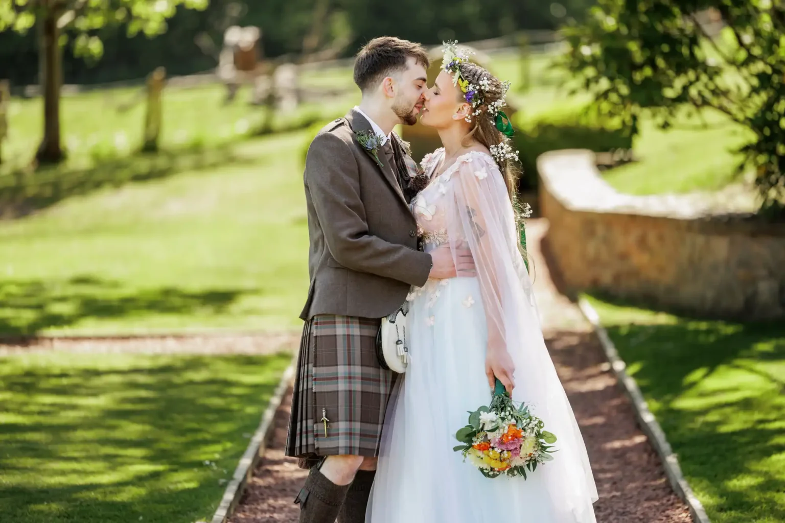 A couple dressed in wedding attire stands on a garden path, sharing a kiss. The groom wears a kilt and the bride holds a bouquet, with greenery and sunlight in the background.