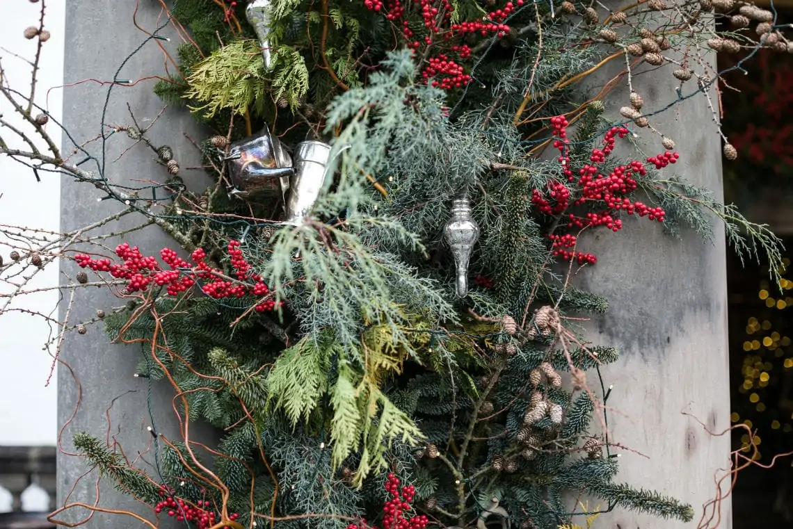 A festive holiday wreath adorned with red berries, pine cones, and silver ornaments attached to a gray concrete column.
