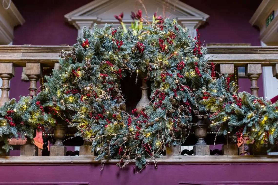 Festive christmas garland with lights and red berries draped over a wooden banister, against a purple wall.
