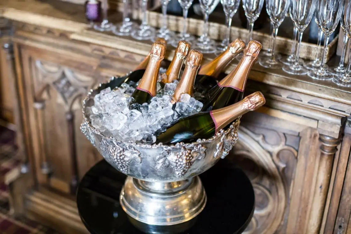 A silver bowl filled with ice and several champagne bottles, next to glasses on a table with a wooden backdrop.