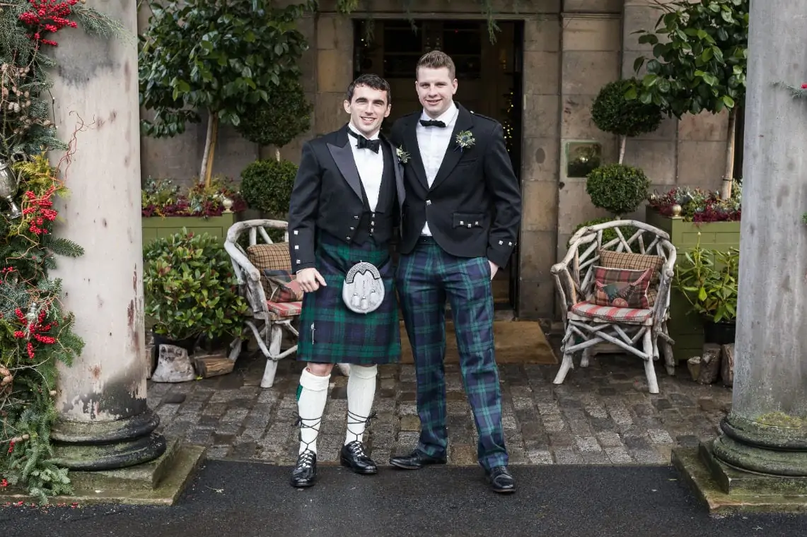 Two men in traditional scottish attire, including kilts and sporran, smiling in front of a building with festive foliage.