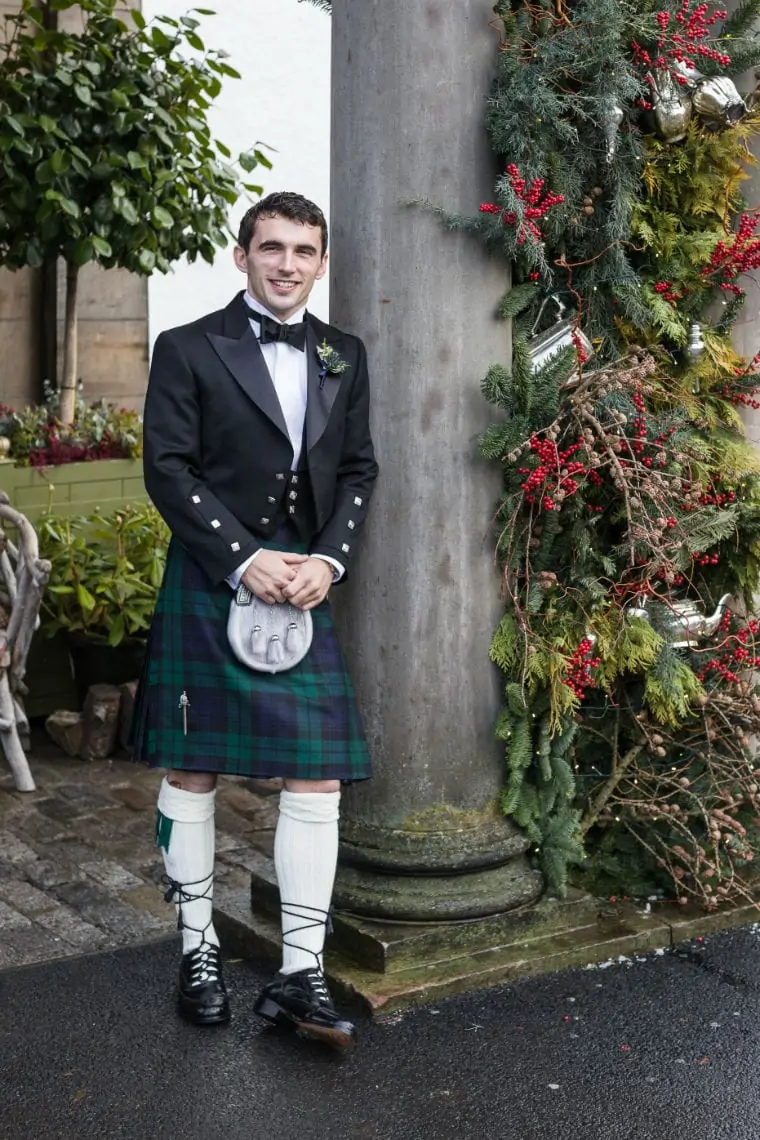 Groom wearing Highland kilt outfit leaning against stone column at the entrance of the hotel