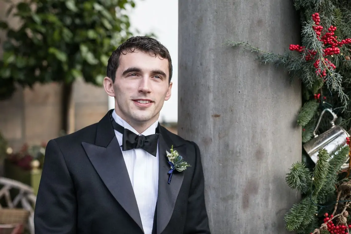 A man in a tuxedo and bow tie smiling, standing next to a column decorated with christmas greenery.