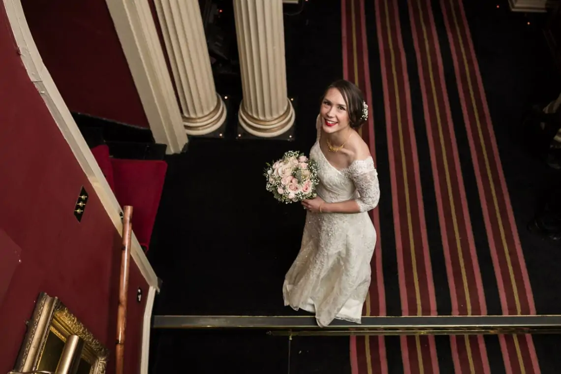 A bride in an off-shoulder wedding dress smiling, holding a bouquet while standing on a staircase with red carpet and white columns.