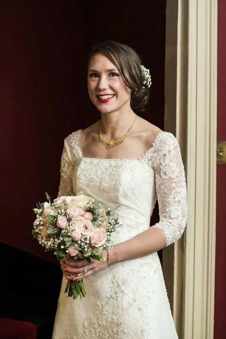 A bride in a lace dress, smiling, holds a bouquet of pink roses and greenery, standing against a red wall.
