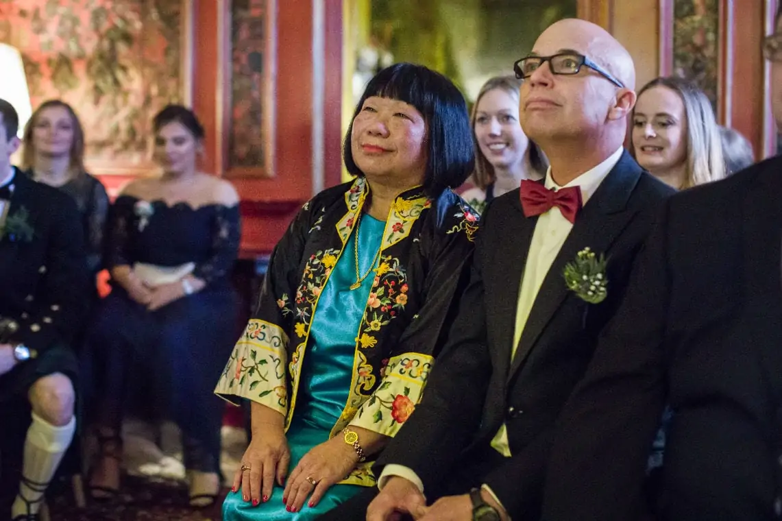 A woman in a colorful traditional dress and a man in a tuxedo with a red bow tie sitting together, smiling and watching an event attentively.