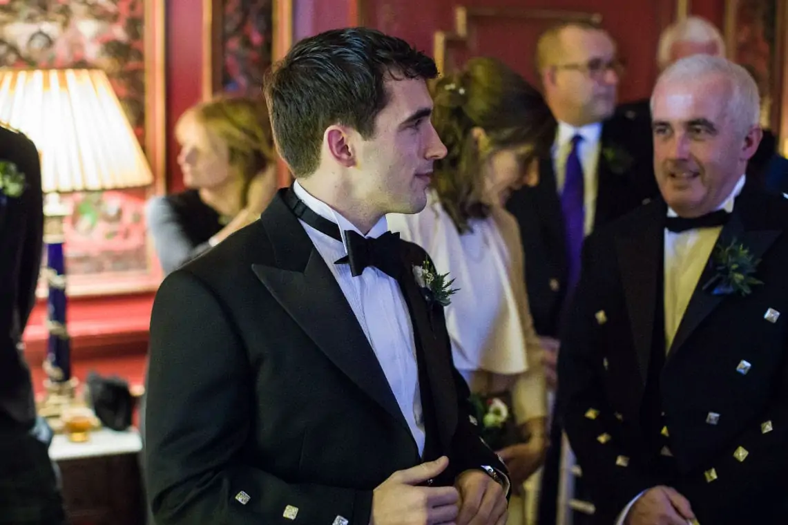 A young man in a tuxedo with a boutonniere at a formal event, conversing with an older man in a traditional kilt outfit, surrounded by guests.