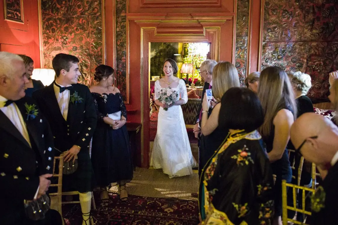 A bride in a white and floral dress smiles while walking through a crowd of guests in a room with red patterned walls.