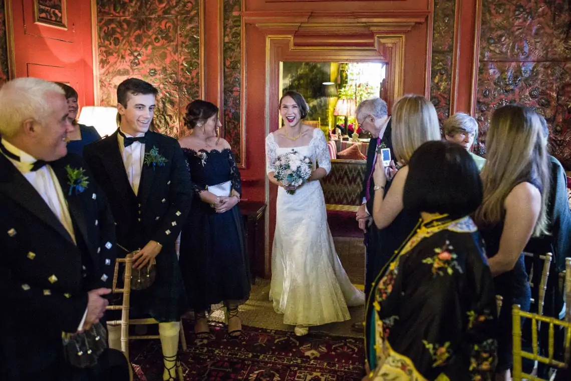 Bride in a white gown smiling in a room with guests dressed in formal attire, including kilts, at a wedding reception.