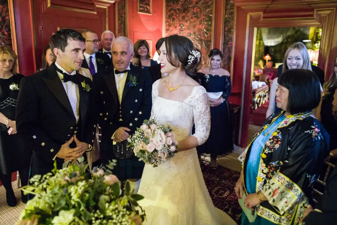 Bride and groom walk through a crowd of guests in an ornately decorated room, smiling as they receive congratulations.