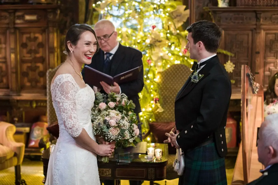 A bride in a lace dress and a groom in a kilt exchanging vows, smiling at each other, with a christmas tree in the background.