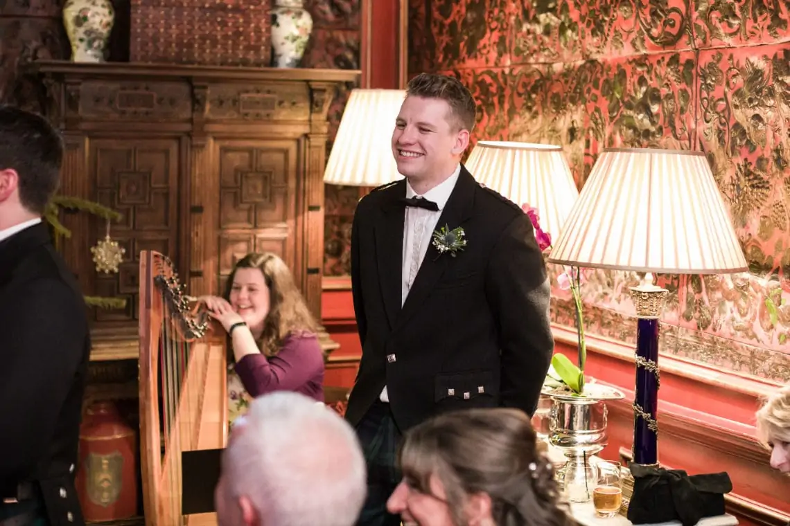 A man in a tuxedo smiling at a formal event in a richly decorated room with vintage furnishings and floral wallpaper.