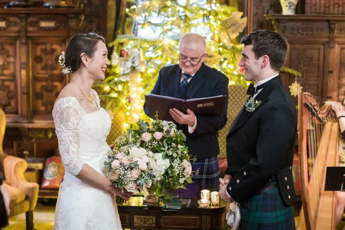 A bride in a white gown and a groom in a kilt smiling at each other during a wedding ceremony, with an officiant and a decorated christmas tree in the background.