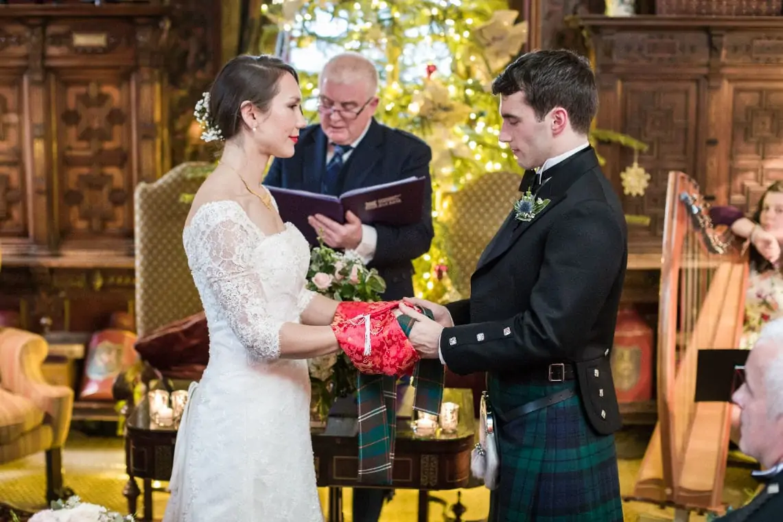 A bride in a lace dress and a groom in a tartan kilt exchange rings at a wedding ceremony, with an officiant and guests in the background.