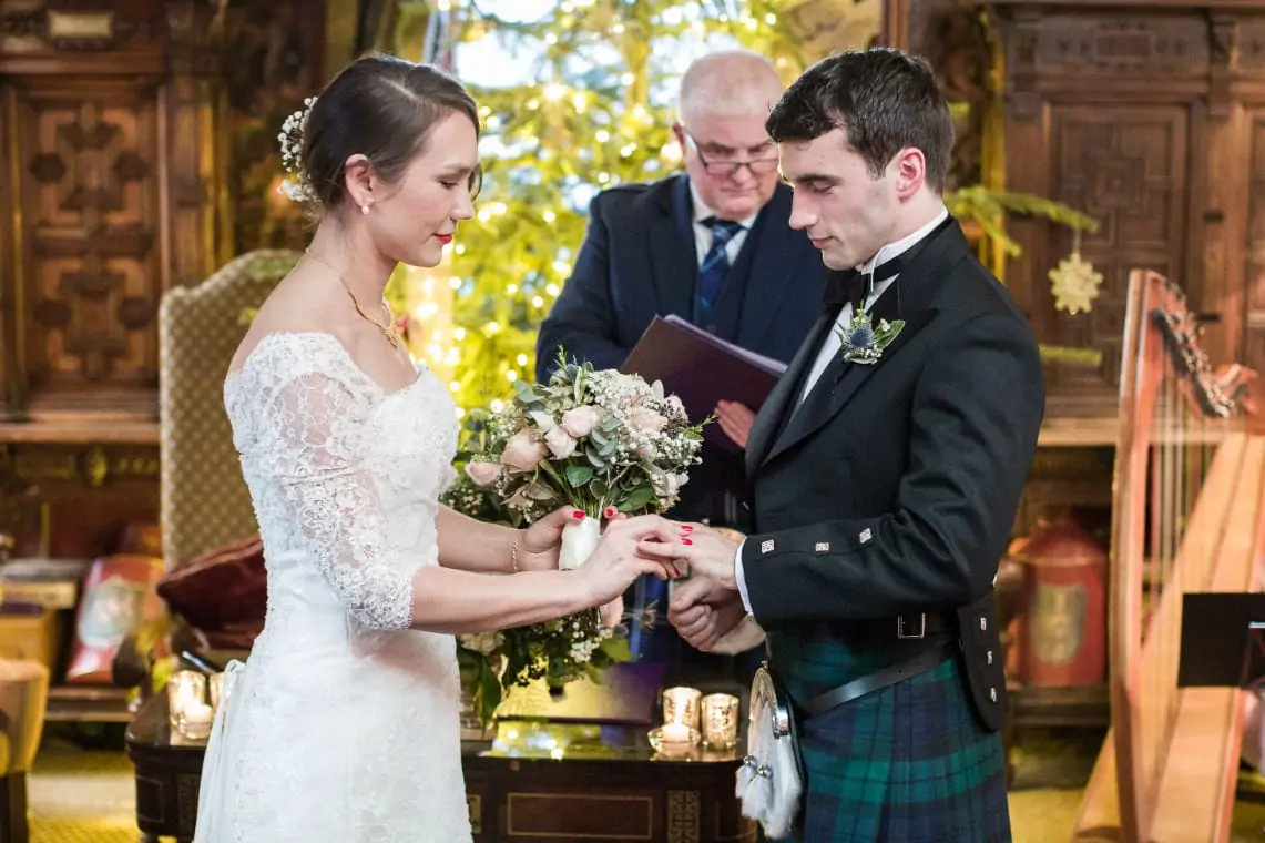 A bride and groom exchange rings during a wedding ceremony in an ornate room, with the groom wearing a traditional kilt.