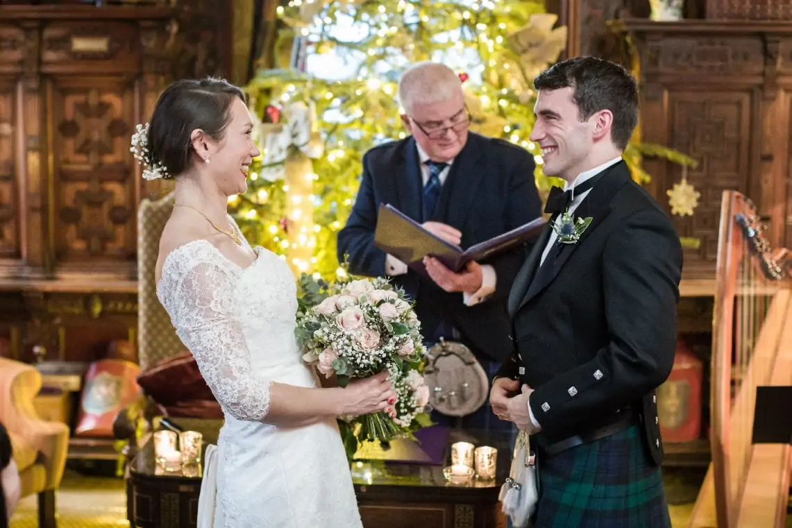 A bride and groom smiling at each other during their wedding ceremony, with the bride holding a bouquet and the groom in a kilt, with an officiant speaking in the background.