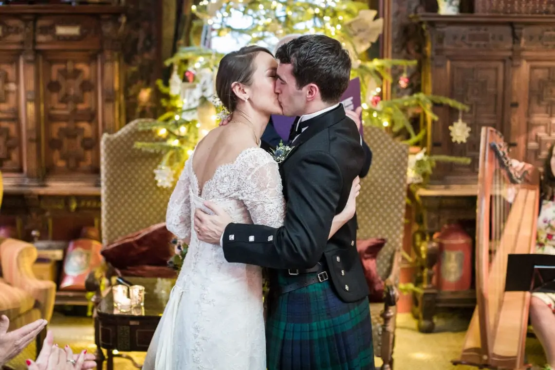 A bride and groom kiss passionately in a grandiose room decorated for christmas, with the bride in a lace dress and the groom in a tartan kilt.