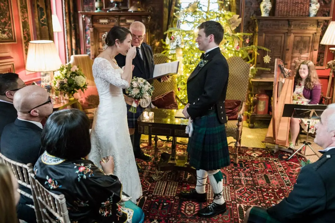 A bride and groom exchange vows in a richly decorated room with an officiant and guests watching, the groom wears a kilt.