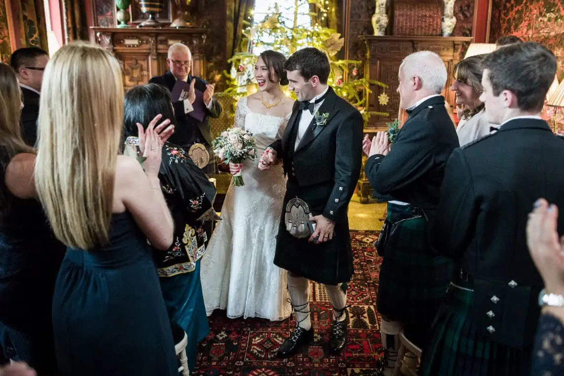 A bride and groom walking down the aisle, smiling as guests applaud; some guests are dressed in traditional scottish kilts.