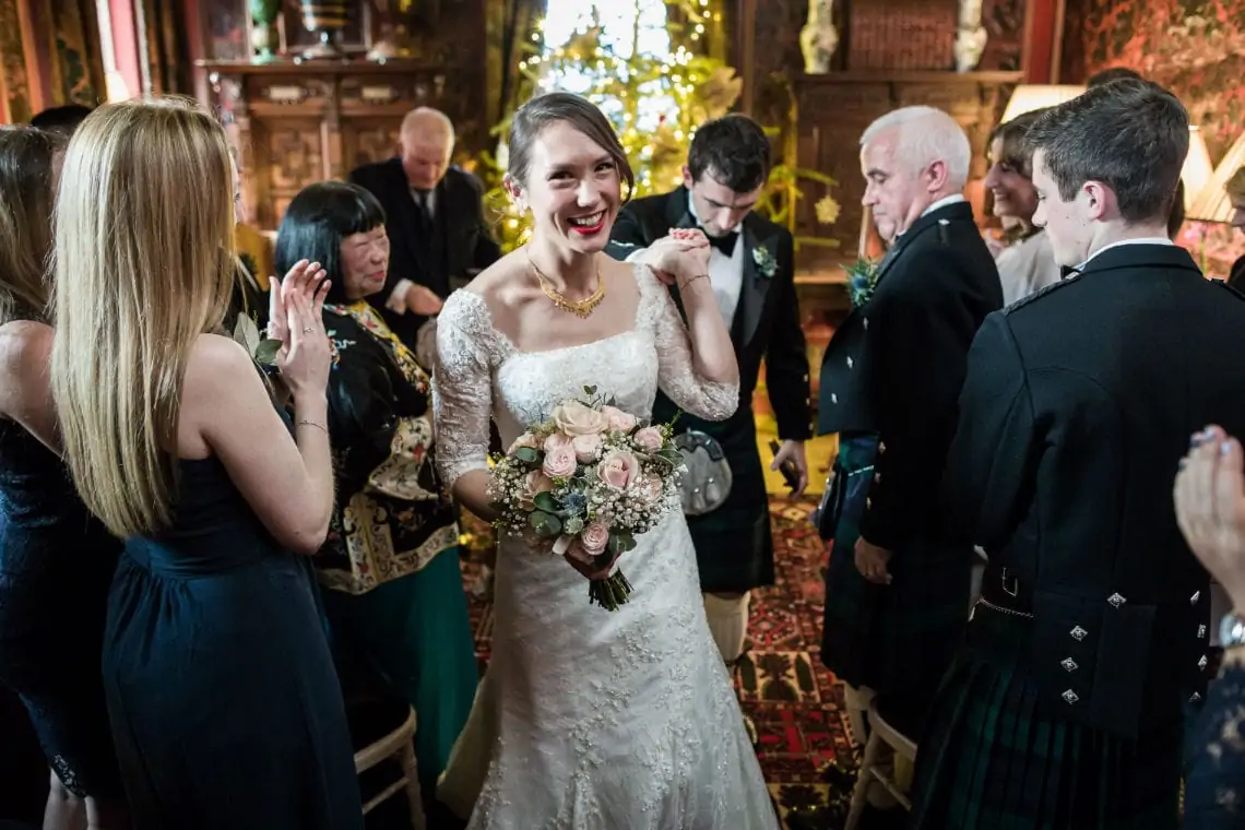 A joyful bride in a lace gown smiling and walking through a clapping crowd in a lavishly decorated room.