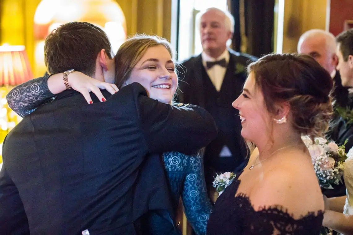 Three people sharing a joyful hug at a formal event, with others in the background. two women are smiling brightly while one is partially obscured.