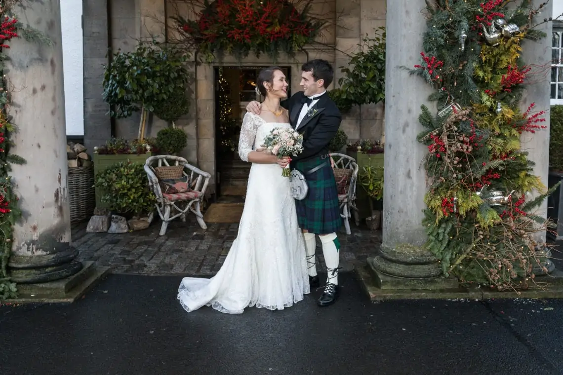 A bride in a white dress and a groom in a kilt embracing in front of a building adorned with christmas decorations.