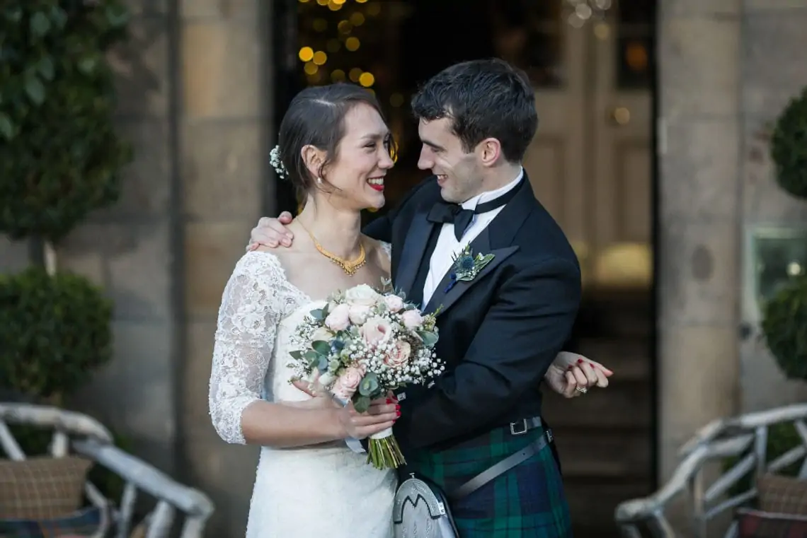 A bride and groom smiling at each other, the bride in a lace dress with a bouquet, and the groom in a tartan kilt, standing outdoors.