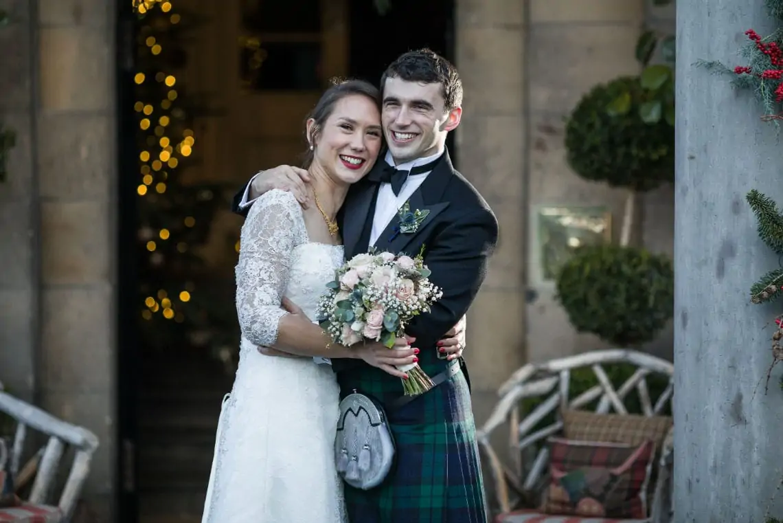 A newlywed couple smiling joyfully, the bride in a white dress holding a bouquet, and the groom in a tartan kilt, outside a festively decorated venue.