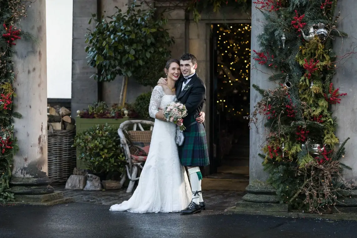 Bride in a white lace gown and groom in a tartan kilt smiling outside a decorated doorway with christmas wreaths and lights.