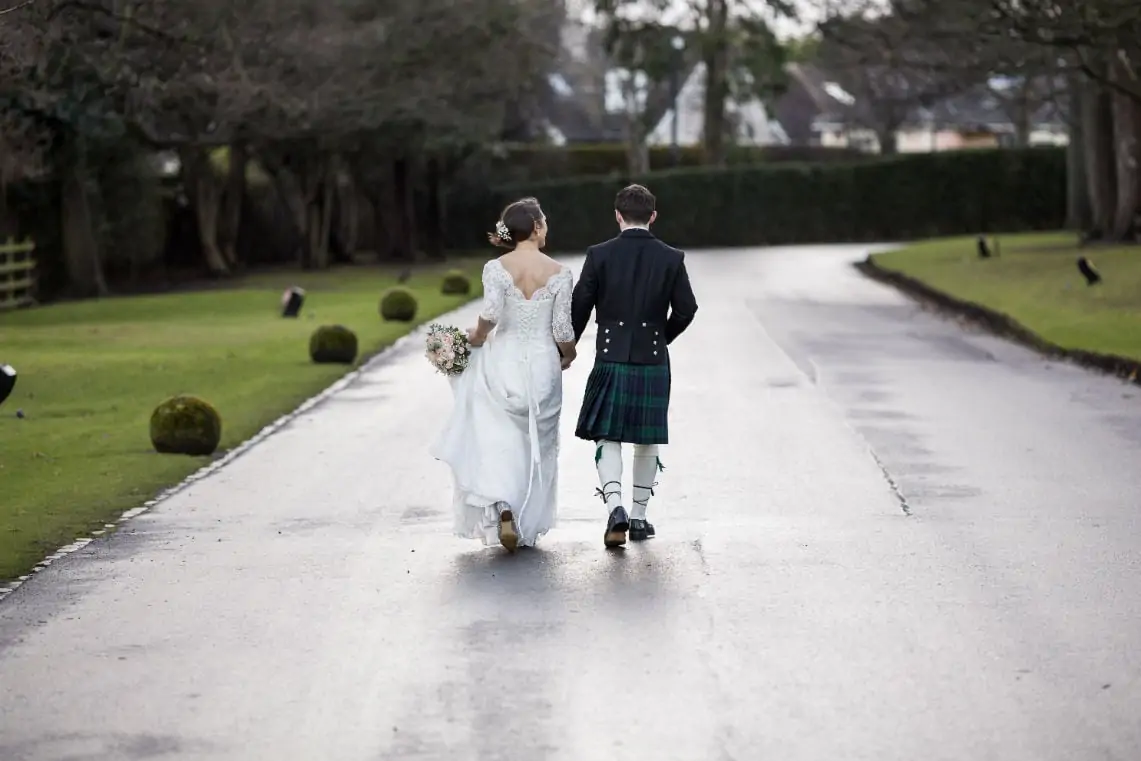 A bride and groom walking down a road, the groom wearing a kilt and the bride in a white dress carrying a bouquet.
