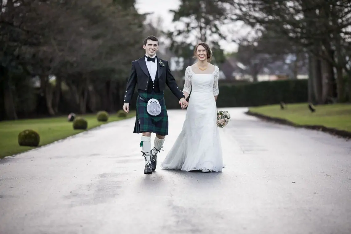 Bride and groom joyfully walking down a pathway, the groom in a kilt and the bride in a white gown, with greenery on the sides.