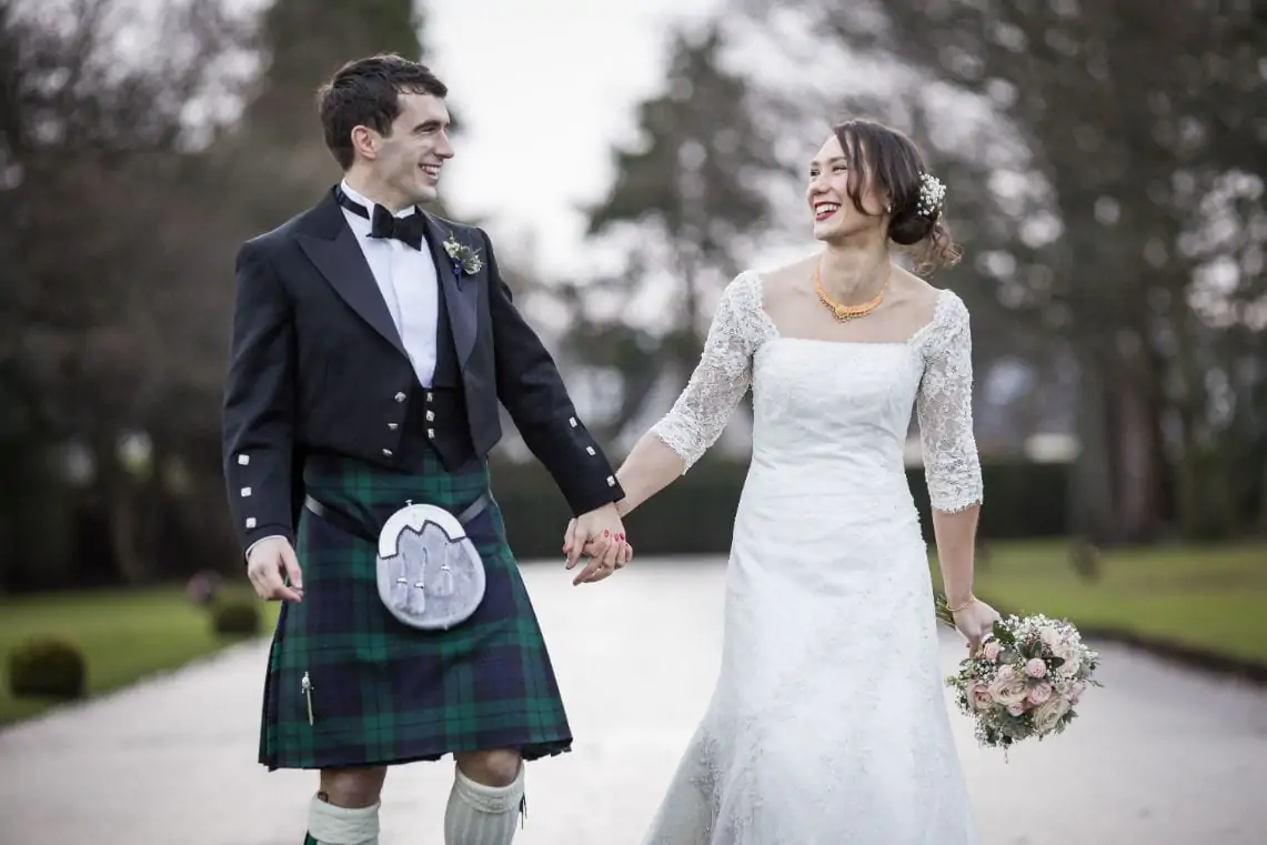 A bride and groom holding hands and smiling at each other, the groom wearing a kilt and the bride in a lace wedding dress, walking through a park.