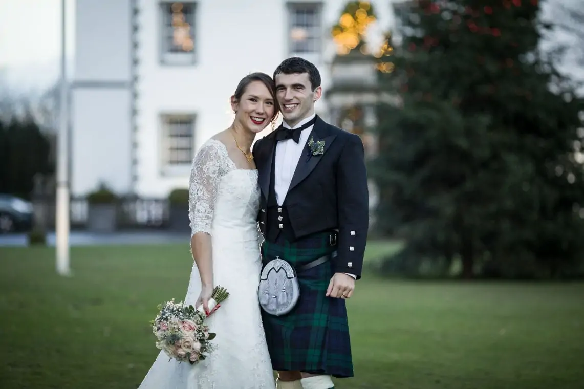 A newlywed couple smiling outside, the bride in a white lace dress with a bouquet, and the groom in a kilt and sporran, with a building and a christmas tree in the background.
