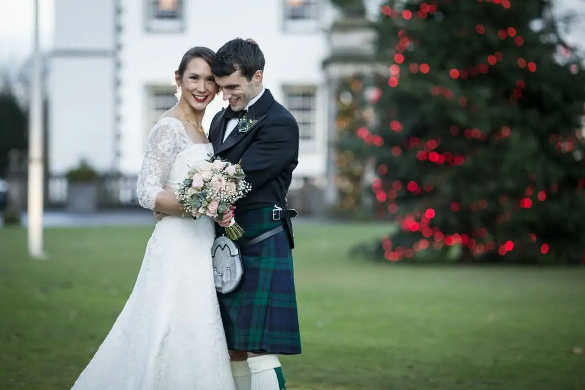A bride in a white dress and a groom in a kilt embrace, smiling, with a bouquet and a christmas tree in the background.