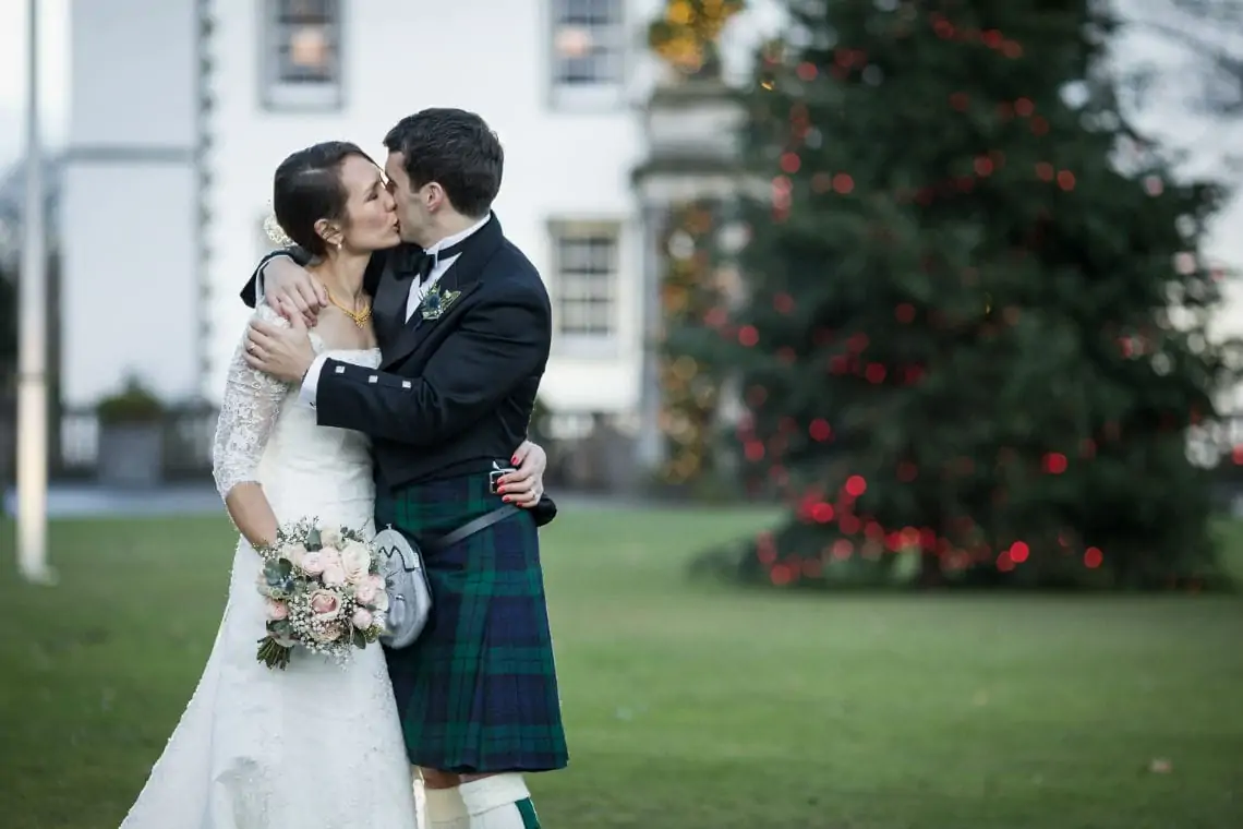 A bride and groom kiss outside, with the groom in a tartan kilt and the bride in a lace dress. a decorated christmas tree is visible in the background.