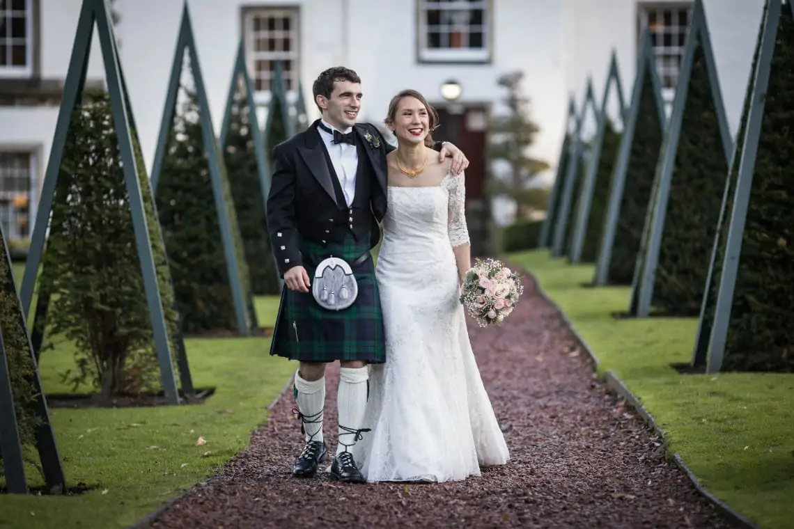A joyful bride and groom, the man in a kilt and the woman in a white gown, walking hand in hand along a garden path.