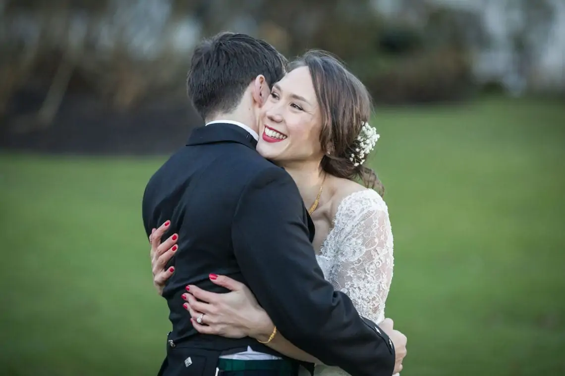 A bride in a lace dress embracing a groom in a black suit and kilt, both smiling joyfully in a grassy field.