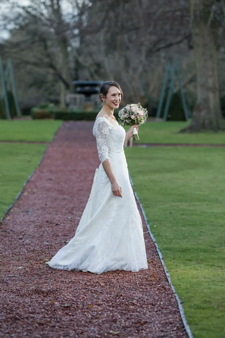 A bride in a lace-sleeved wedding dress smiles while holding a bouquet, walking down a tree-lined garden path.