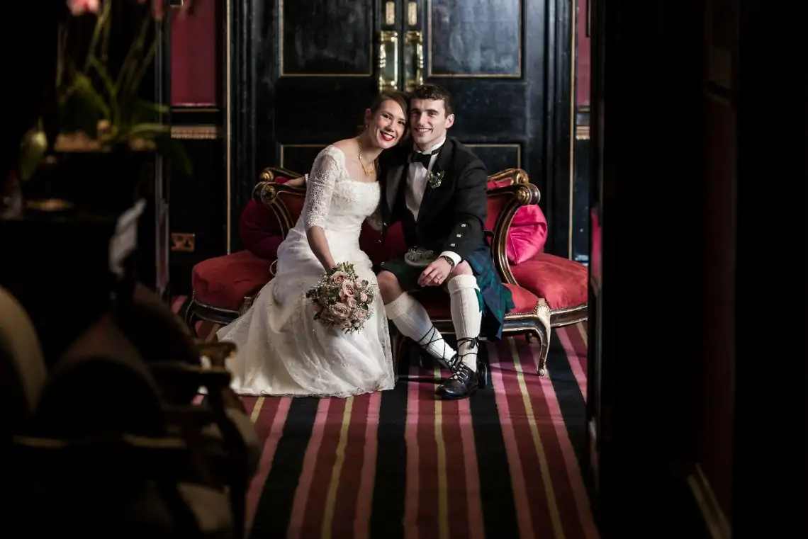 A newlywed couple smiling and sitting together on a red velvet couch in a luxurious room with striped carpet and dark wood doors.