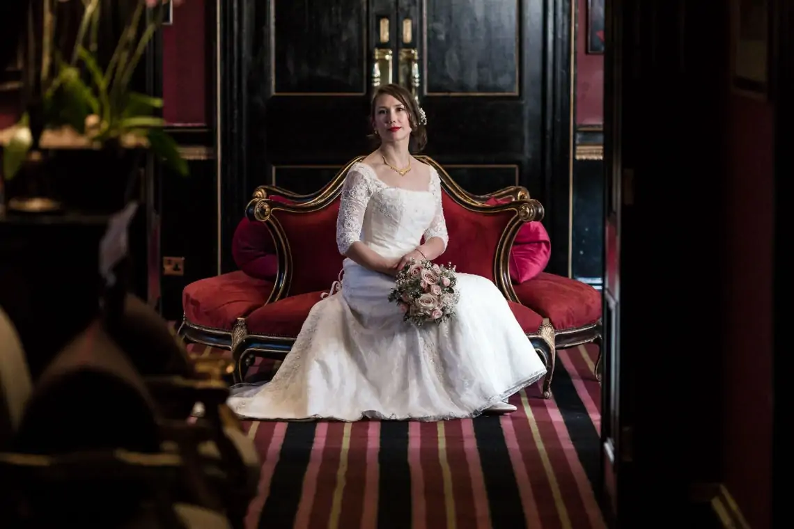 A bride in a white lace gown sits elegantly on a red velvet sofa, holding a bouquet, in a richly decorated room with dark walls and striped carpet.