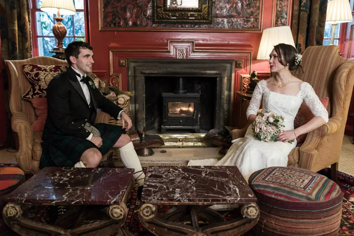 A newlywed couple, a man in a kilt and a woman in a vintage wedding dress, sitting in a cozy room with a lit fireplace, smiling at each other.