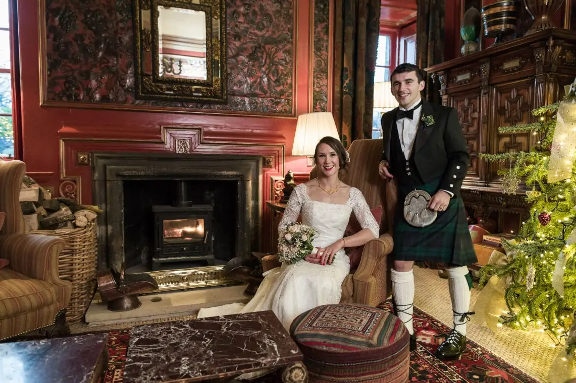 A bride in a white dress and a groom in a kilt smile by a fireplace in an opulent room decorated for christmas.