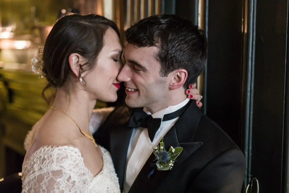 A bride in a lace dress and groom in a tuxedo sharing a close, affectionate moment, smiling at each other indoors.