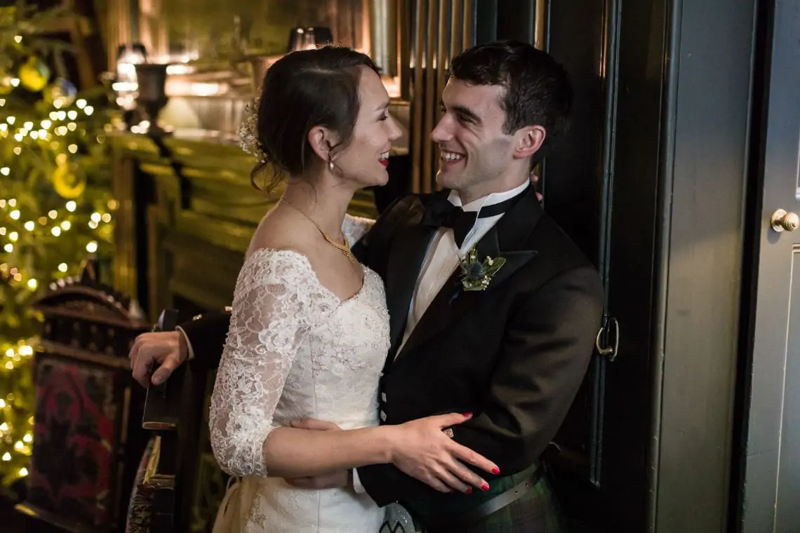 A bride in a lace dress and a groom in a kilt embracing and smiling at each other in a dimly lit room with fairy lights.