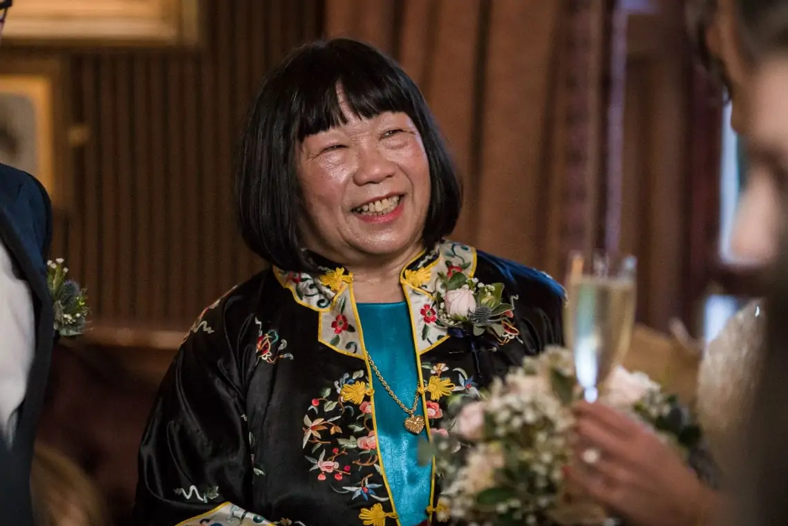 A joyful elderly asian woman in a floral embroidered traditional dress, smiling at a wedding reception while holding a champagne glass.