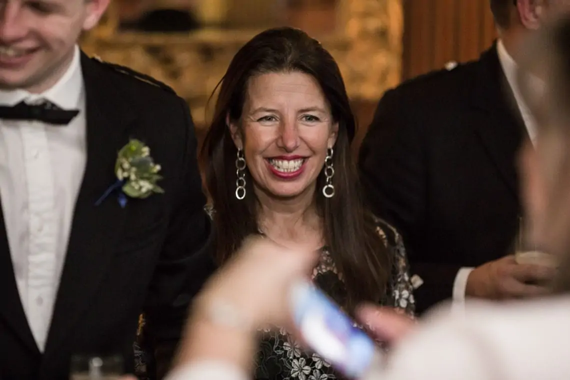A woman with long brown hair, wearing earrings and a black floral dress, smiling at a wedding reception. a man in a tuxedo is partially visible on the left.