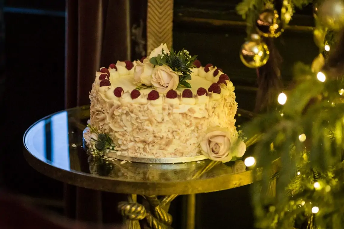 A decorated cake with white frosting, topped with raspberries and roses, is displayed on a mirrored table beside a lit christmas tree.