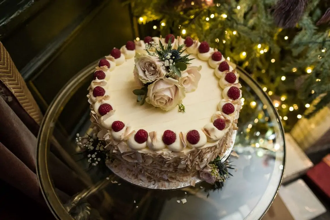 A decorated cake with raspberries, pink roses, and coconut flakes on a silver platter, set against a christmas tree backdrop.
